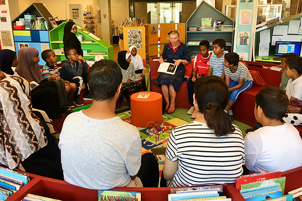 Børn og voksne siddende i rundkreds på bibliotek med en fortæller, som læser op af en bog