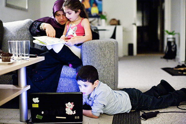 Mor hjælper datter med lektier, mens mindre søn kigger på en pc