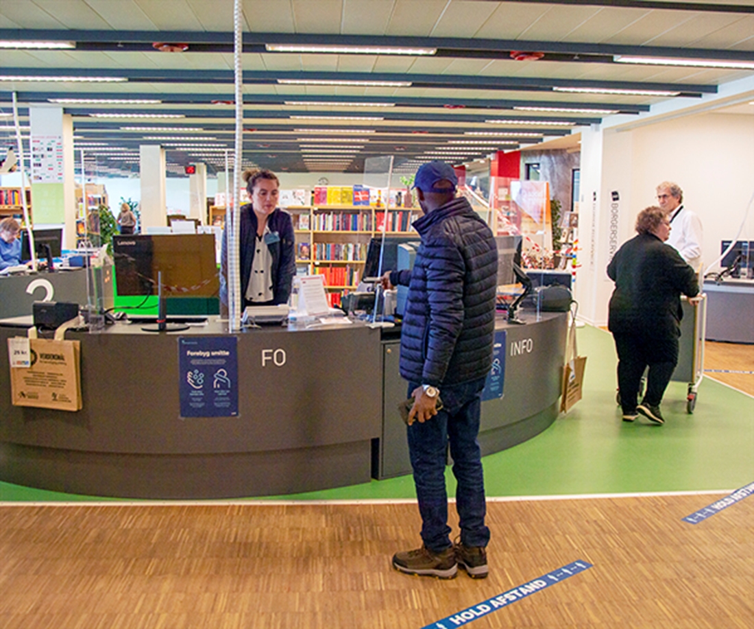 Info-skranke på bibliotek, hvor kvindelig bibliotekar vejleder en herre