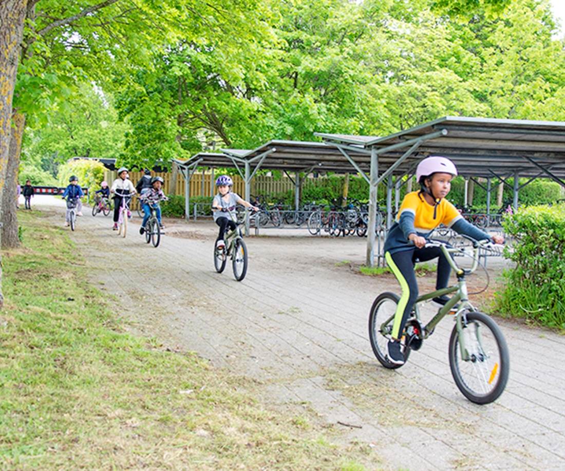 Børn i 8-års alderen iført cykelhjelme cykler forbi cykelparkering ved skole